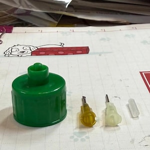 May include: A green plastic bottle cap and three small, clear, and yellow plastic components. The components appear to be parts of a dispenser or applicator. The items are on a white surface with a dog cartoon.