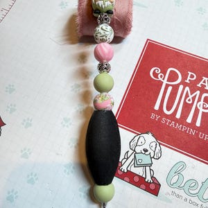 May include: A decorative pin featuring a gnome bead, floral beads, and a black oval bead. The pin has a silver top and a sharp, pointed end. The beads are in shades of green, pink, and white.