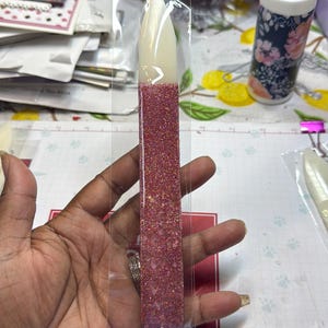 May include: A clear plastic package containing a nail art tool. The tool has a white tip and a pink glitter-filled body. The nail art tool is held in a hand.