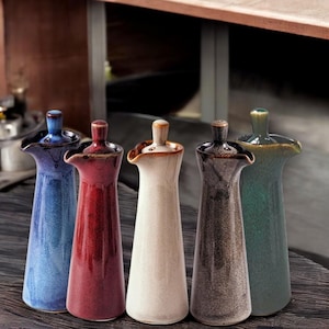May include: Five ceramic oil and vinegar bottles with pour spouts and lids. The bottles are in various colours, including blue, red, white, grey, and green.