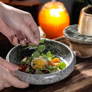 Op de afbeelding: Een grijze gespikkelde keramische kom gevuld met een salade van verse groenten, groenten en een gehalveerd hardgekookt ei. De kom staat op een houten oppervlak.