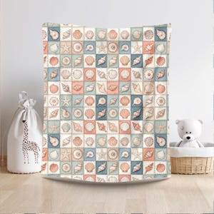 Puede incluir: Una manta estampada cuadrada con un dise&ntilde;o repetido de conchas marinas y estrellas de mar en tonos melocot&oacute;n, azul y crema. La manta se muestra sobre un fondo neutro, lo que sugiere un tema costero o de playa.