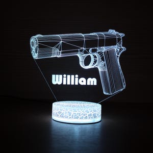 以下が含まれることがあります： 白い文字で「William」と書かれた、銃のデザインの3Dプリントのナイトライト。銃はワイヤーフレーム効果で描かれ、ベースはテクスチャパターンになっています。光は柔らかく光ります。