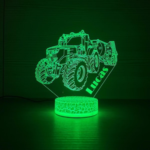 Puede incluir: Una luz nocturna LED verde con forma de tractor con el nombre "Lucas" grabado en la base. El tractor es un contorno detallado, y la base tiene un diseño de tierra agrietada. La luz está enchufada.