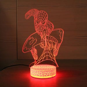 May include: A red Spider-Man 3D LED lamp. The lamp features a detailed outline of the superhero, with a web design. The base of the lamp is round and has a cracked pattern, emitting a warm red glow. The lamp is a decorative item.