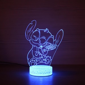 Puede incluir: Una luz nocturna LED azul que representa al personaje de dibujos animados Stitch sosteniendo una muñeca. La luz tiene una base con efecto de vidrio agrietado y un contorno brillante del personaje. La luz está enchufada a una fuente de alimentación.