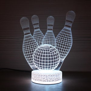 Puede incluir: Una lámpara de noche blanca e iluminada con temática de bolos. El diseño presenta una bola de bolos y bolos con un patrón de cuadrícula. La luz se encuentra sobre una base redonda con un patrón decorativo. La lámpara está enchufada a una toma de corriente.