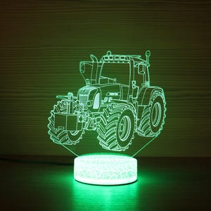 Può includere: Una lampada a illusione ottica 3D che presenta il contorno luminoso verde brillante di un trattore. Il design del trattore è inciso su un pannello acrilico trasparente posto su una base circolare illuminata che emette luce verde.