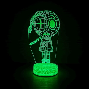 Könnte beinhalten: Eine grüne LED-Lampe mit einem Drahtgitter-Cartoon-Charakter-Design. Die Figur hat ein Kreuz als Auge und das Wort "Marry" steht auf der Seite. Die Lampe steht auf einem runden Sockel mit einem strukturierten Muster.