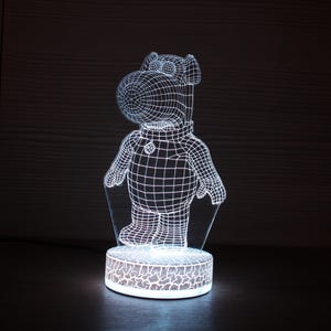 Może przedstawiać: Nocna lampka z efektem 3D przedstawiająca postać hipopotama z kreskówki. Hipopotam jest przedstawiony w białym stylu drucianym, stojąc na okrągłej podstawie z wzorem. Światło emituje delikatną poświatę, tworząc unikalny element dekoracyjny.