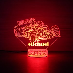 Pode incluir: Uma luz noturna LED vermelha com um design de carro de corrida de Fórmula 1. O nome "Michael" é exibido abaixo do carro. A base tem um padrão rachado e a luz emite um brilho vermelho.
