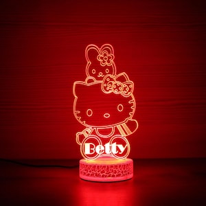 Puede incluir: Una luz de noche LED roja con un personaje de dibujos animados con un lazo y un conejito. El nombre "Betty" se muestra en la base. La luz está sobre una superficie oscura con un fondo rojo.