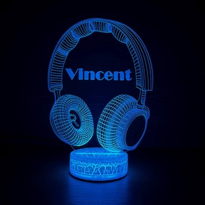 Op de afbeelding: Een blauw LED-nachtlampje in de vorm van een koptelefoon, met de naam "Vincent" weergegeven. De koptelefoon heeft een draadframe-ontwerp en de basis heeft een craquelé-effect. Het licht straalt een koele blauwe gloed uit.