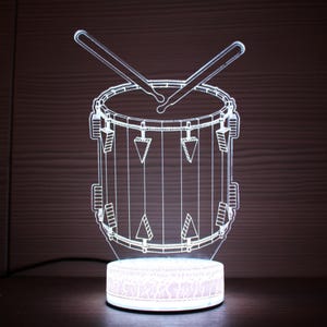 Puede incluir: Una luz nocturna de acrílico blanco con forma de tambor con dos baquetas. El tambor tiene un diseño detallado con líneas y triángulos.