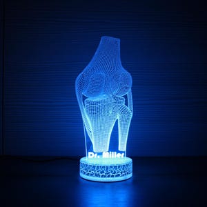 Op de afbeelding: Een blauw verlicht 3D-model van een kniegewricht, met een draadframe ontwerp. Het model wordt verlicht met een zacht blauw licht en staat op een voet met de tekst "Dr. Miller". De voet heeft een craquelé patroon.