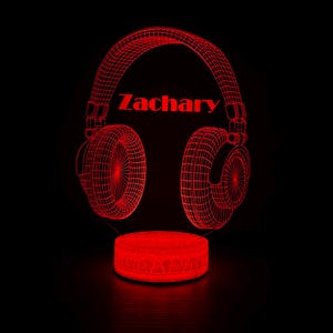 Puede incluir: Un diseño de auriculares rojo renderizado en 3D con el nombre "Zachary" en texto rojo. Los auriculares están iluminados y parecen estar hechos de un diseño de estructura alámbrica. La base también es roja e iluminada, sobre un fondo negro.