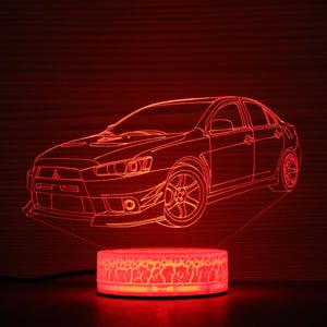 Puede incluir: Una luz nocturna LED roja con forma de coche Mitsubishi Lancer Evolution. La luz está encendida e ilumina el contorno del coche.
