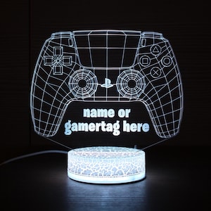 May include: A glowing white 3D LED lamp shaped like a video game controller. The controller design features a grid pattern and the text "name or gamertag here". The lamp sits on a textured, illuminated base.