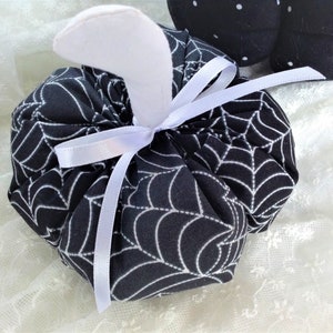 May include: A black and white fabric pumpkin with a white spiderweb pattern and a white satin ribbon tied around the stem.