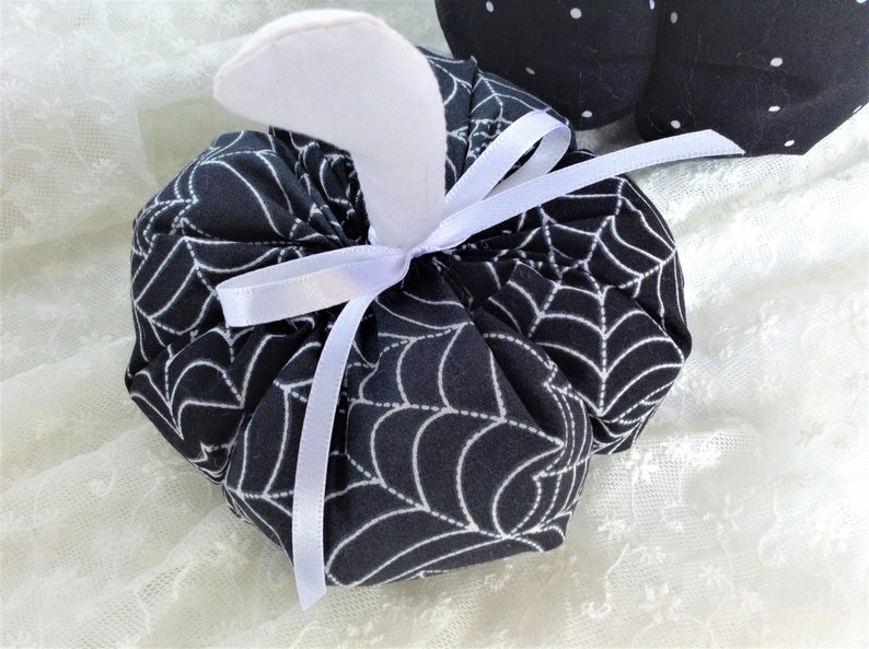 May include: A black and white fabric pumpkin with a white spiderweb pattern and a white satin ribbon tied around the stem.