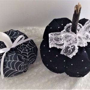 May include: Two black fabric pumpkins, one with a white spiderweb pattern and a white satin ribbon, the other with white polka dots and a white lace bow.