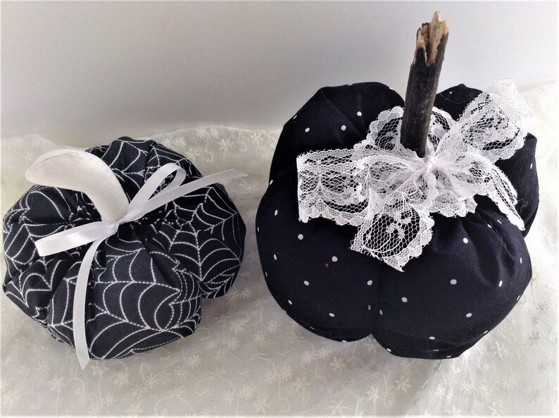 May include: Two black fabric pumpkins, one with a white spiderweb pattern and a white satin ribbon, the other with white polka dots and a white lace bow.