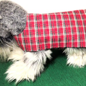 May include: A plush dog toy with a red and green plaid coat. The dog has gray fur on its head and tail, and white and gray fur on its legs. The coat is trimmed with black fabric.