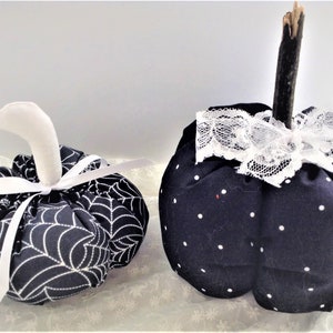 May include: Two black fabric pumpkins with white accents. One pumpkin has a white spiderweb pattern and a white ribbon. The other pumpkin has white polka dots and a white lace bow.