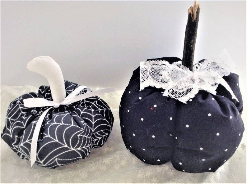 May include: Two black fabric pumpkins with white accents. One pumpkin has a white spiderweb pattern and a white ribbon. The other pumpkin has white polka dots and a white lace bow.