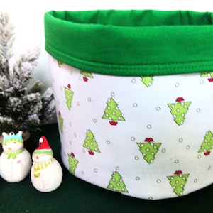 May include: A white fabric storage basket with a green rim and a pattern of green Christmas trees with red accents. Three snowman figurines and a small Christmas tree are in the background.