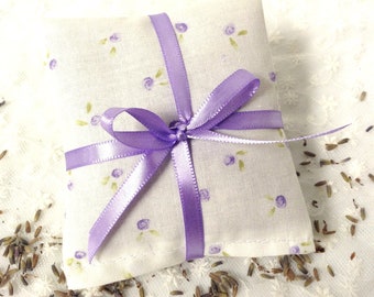 May include: A square fabric sachet featuring a white background with a small purple floral and green leaf pattern. The sachet is tied with a smooth, shiny purple satin ribbon that is knotted in a bow across the center of the square textile item.