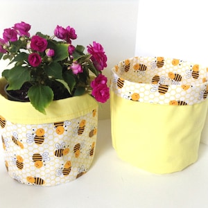 May include: Two yellow fabric plant pots. One pot holds a plant with magenta flowers. The pots have a yellow exterior and a bee and honeycomb pattern on the interior trim. The other pot is empty.
