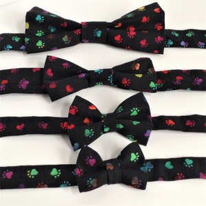 May include: Four black bow ties with colorful paw print patterns. The bow ties are arranged in a row, each with a different size. The paw prints are in shades of red, blue, green, and yellow.