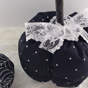 May include: Two black and white fabric pumpkins. One pumpkin has white polka dots and a white lace bow. The other pumpkin has a white spiderweb design.