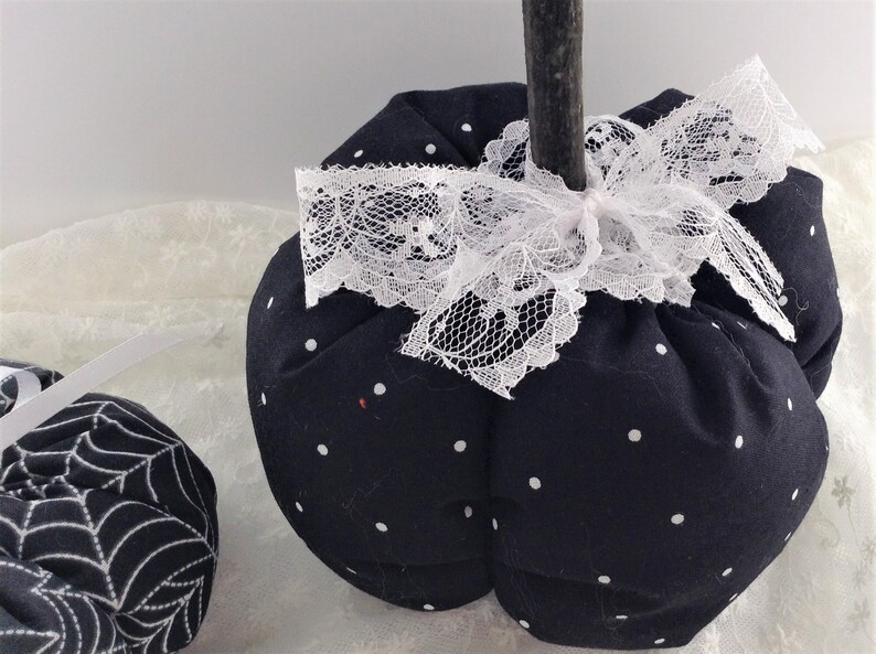 May include: Two black and white fabric pumpkins. One pumpkin has white polka dots and a white lace bow. The other pumpkin has a white spiderweb design.