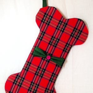 May include: A Christmas stocking in the shape of a dog bone, made of red plaid fabric with black and green accents. A green bow with a pink button is attached. The stocking hangs from a candy cane.