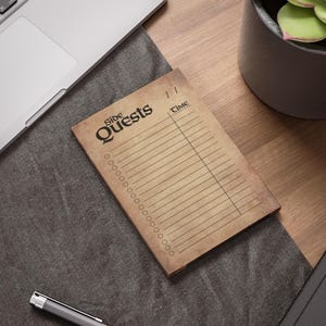 May include: A notepad with the words "Side Quests" in a vintage font. The notepad has a lined section for notes and a section for time. The notepad is on a wooden desk next to a laptop and a pen.