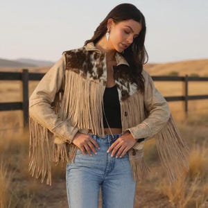 Könnte beinhalten: Beigefarbene Wildlederjacke mit Fransen und Kuhfell-Akzenten auf den Schultern. Die Jacke hat eine Knopfleiste und einen klassischen Western-Stil. Das Model trägt eine blaue Jeans und ein schwarzes Top.