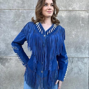 Könnte beinhalten: Blaue Wildlederjacke mit langen Fransen. Die Jacke hat weiße Ziernähte am Kragen, an den Ärmeln und an der Vorderseite. Die Jacke ist zugeknöpft und wird mit einer blauen Jeans getragen.