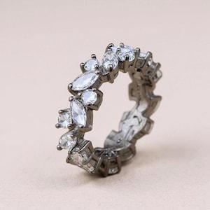 Pode incluir: Um anel prateado com um padrão contínuo de pedras preciosas claras e brilhantes. O anel apresenta uma banda de metal escuro com um design texturizado. As pedras são de várias formas.