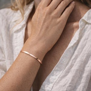 Può includere: Un bracciale a polsino in oro rosa. Il bracciale è una semplice fascia sottile con una finitura liscia e lucida. Il gioiello è indossato sul polso di una persona che indossa una camicia bianca abbottonata.