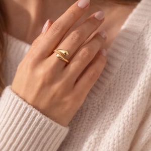 Pode incluir: Um anel dourado com um design único e fluido. O anel envolve o dedo com duas extremidades arredondadas que se encontram. A joia é exibida em uma mão, mostrando a forma elegante e o acabamento polido do anel.