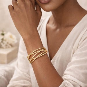 Könnte beinhalten: Ein goldfarbenes Armreif-Armband mit einem verwobenen Design. Das Armband wird am Handgelenk einer Person getragen, und die Goldfarbe kontrastiert mit dem Hautton. Das Armband scheint aus dünnen, gebogenen Metallbändern zu bestehen.