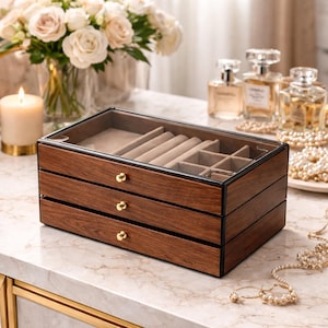 Może przedstawiać: Trzypoziomowe drewniane pudełko na biżuterię ze szklanym blatem. Pudełko ma ciemnobrązowe wykończenie ze złotymi gałkami. Górna szuflada jest otwarta, odsłaniając beżowe wnętrze z przegródkami do przechowywania biżuterii. Pudełko stoi na marmurowej powierzchni.