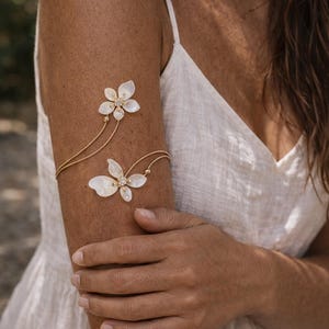 May include: A delicate gold arm cuff featuring two flower-shaped adornments. The upper flower is positioned near the shoulder, while the lower one rests on the arm. Each flower is embellished with small, sparkling accents. The cuff is connected by fine gold chains.