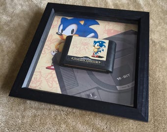 Può includere: Opera d'arte incorniciata con una cartuccia Mega Drive di Sonic the Hedgehog. La cornice nera contiene una stampa con Sonic e una grafica della console Sega. La cartuccia è nera con le parole "MEGA DRIVE" e un'immagine di Sonic. L'opera è esposta su una superficie beige.