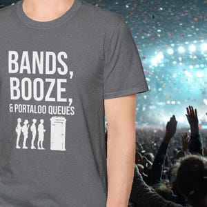 Puede incluir: Una camiseta gris oscuro con el texto "Bands, Booze, & Portaloos Queues" en blanco. La camiseta presenta un gráfico de tres personas de pie en la fila frente a un inodoro portátil.