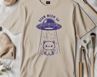 May include: Beige t-shirt with a blue graphic of a UFO beaming up a cartoon cat. The text "BEAM MEOW UP" is above the UFO. The shirt is surrounded by makeup brushes.
