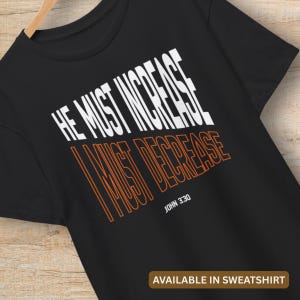 Può includere: T-shirt nero con la scritta "HE MUST INCREASE, I MUST DECREASE" in lettere bianche e arancioni. Il testo "JOHN 3:30" è in basso. La parte inferiore dell'immagine dice "AVAILABLE IN SWEATSHIRT".
