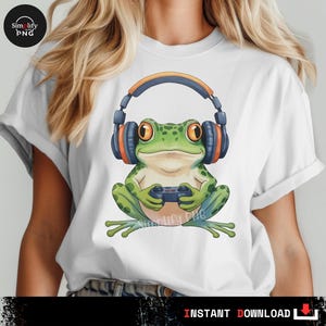 May include: White t-shirt featuring a cartoon frog wearing headphones and holding a game controller. The frog is green with orange eyes and is sitting. The headphones are blue and orange. The words "INSTANT DOWNLOAD" are at the bottom.